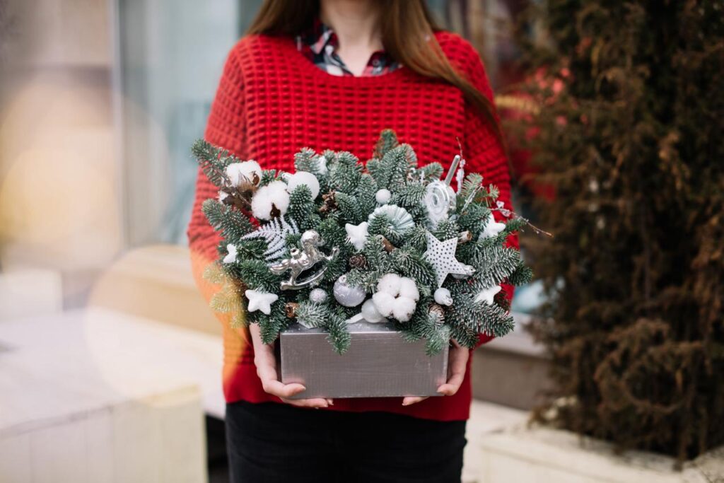Person in a red sweater holding a festive arrangement of pine branches decorated with white ornaments, cotton sprigs, and silver accents, in a silver box.