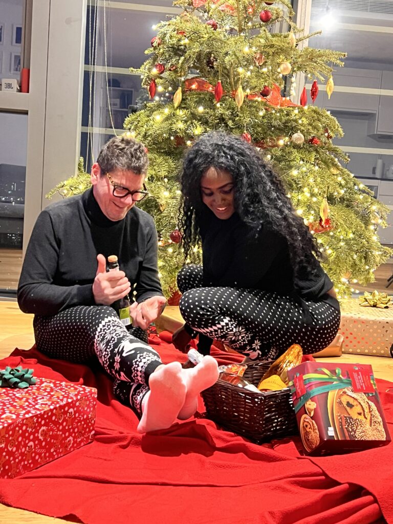 A couple sits on a red blanket in front of a decorated Christmas tree, wearing matching black and white pyjamas. The man holds a bottle while the woman looks into a gift basket. Wrapped presents are under the illuminated tree.