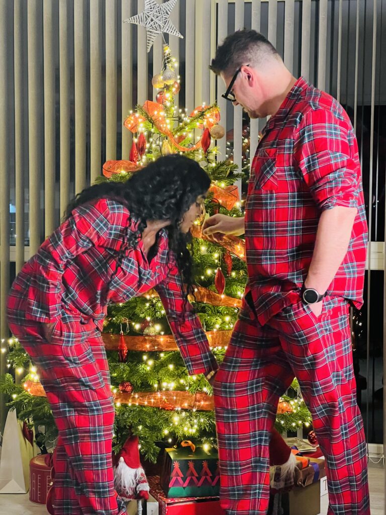 A man and woman in matching red plaid pajamas decorate a Christmas tree with white lights, gold and red ornaments, and a silver star on top.