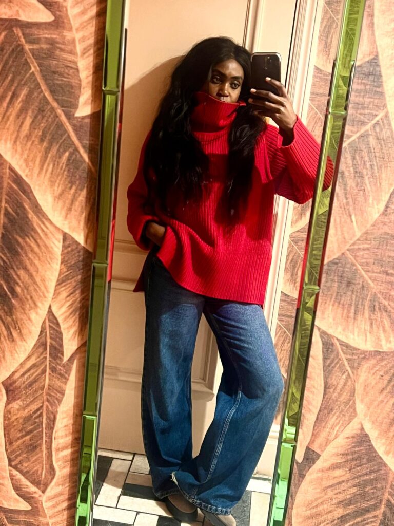 A woman wearing a red chunky turtleneck sweater styled with dark blue jeans, standing in a cozy, warmly lit room taking a selfie.