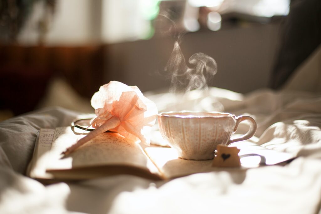 Hot drink, book, glasses, and a small wooden heart on a bed, suggesting a cozy night in alone for those single on Valentine's Day.