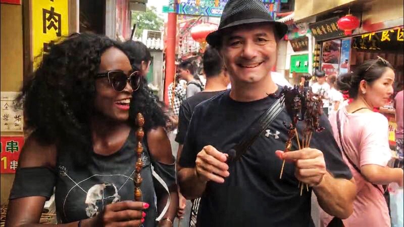 Man and woman gleefully walking through a food market in China while holding grilled insect skewers.