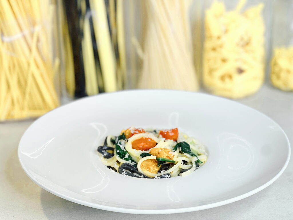 A plated serving of the Meghan Markle one-pot pasta recipe, featuring spaghetti with spinach, red and yellow Datterini tomatoes, and grated cheese.