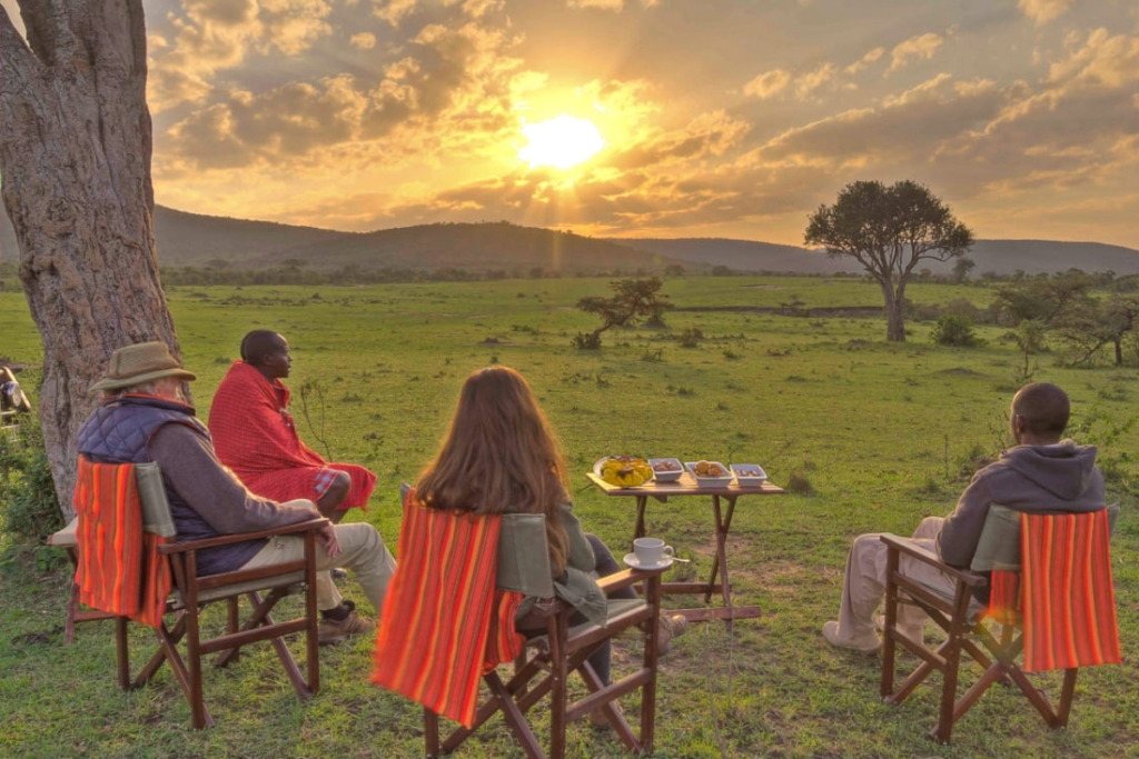 On an African safari, one Maasai guide wears a red shuka while two tourists and another guide have theirs draped over safari chairs, watching the golden savanna sunset
