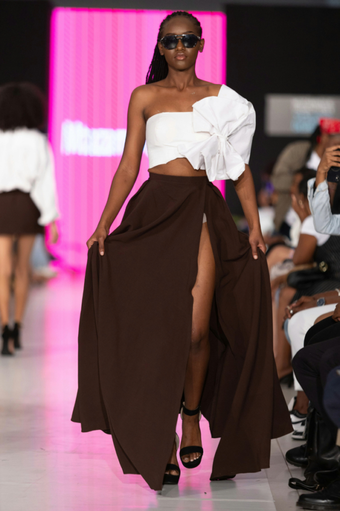 Model walks the runway in a white off-shoulder crop top, brown flowy high-slit skirt, and black sandals.