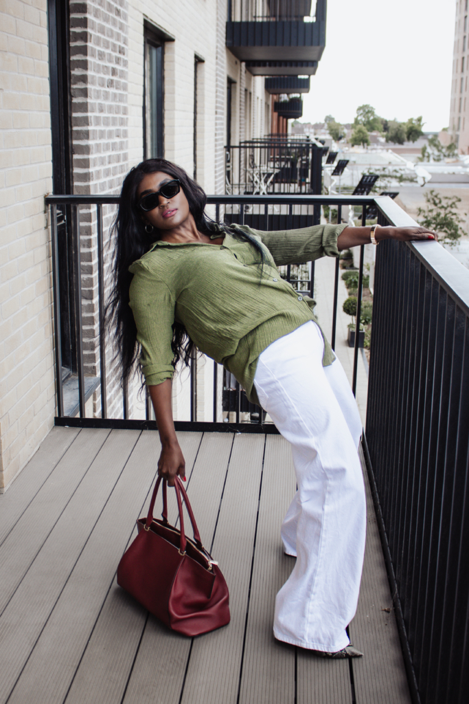 A woman in an olive green shirt and white wide-leg pants leans over a balcony railing, holding a maroon handbag.