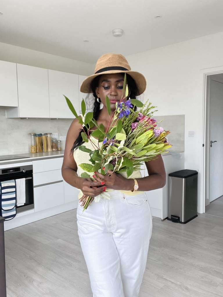 A black woman in white jeans, a white tank top, and a hat stands inside her flat, holding a bouquet of flowers.