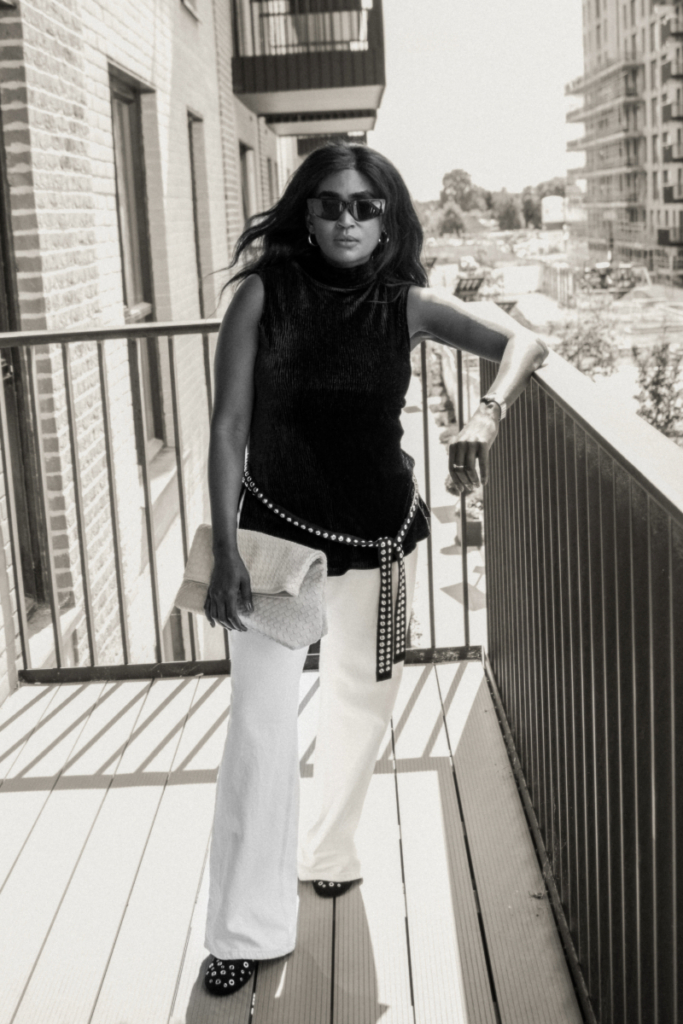 A black and white photo of a woman in a black top, white wide-leg pants, and a chain belt, holding a clutch on a balcony.