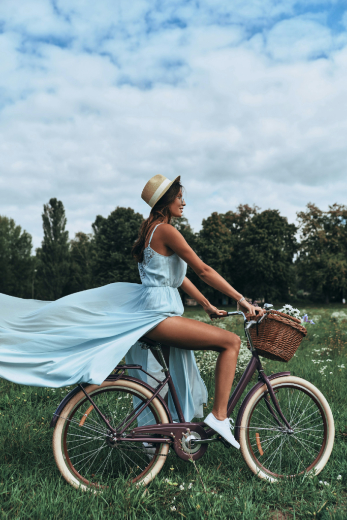 Woman in a baby blue stylish flowy maxi dress and straw hat riding a bicycle, with the wind blowing her dress elegantly behind her.