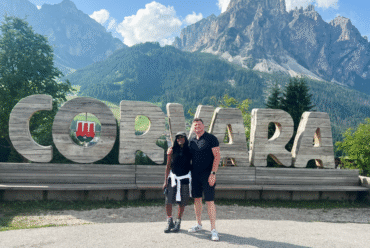 Couple standing in front of the large wooden Corvara sign in the Dolomites, with mountain peaks in the background, showcasing one of the top reasons to visit Corvara in summer.