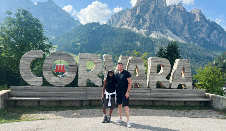 Couple standing in front of the large wooden Corvara sign in the Dolomites, with mountain peaks in the background, showcasing one of the top reasons to visit Corvara in summer.