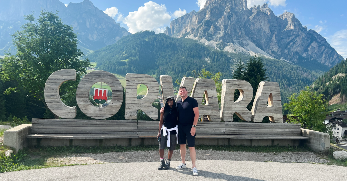 Couple standing in front of the large wooden Corvara sign in the Dolomites, with mountain peaks in the background, showcasing one of the top reasons to visit Corvara in summer.