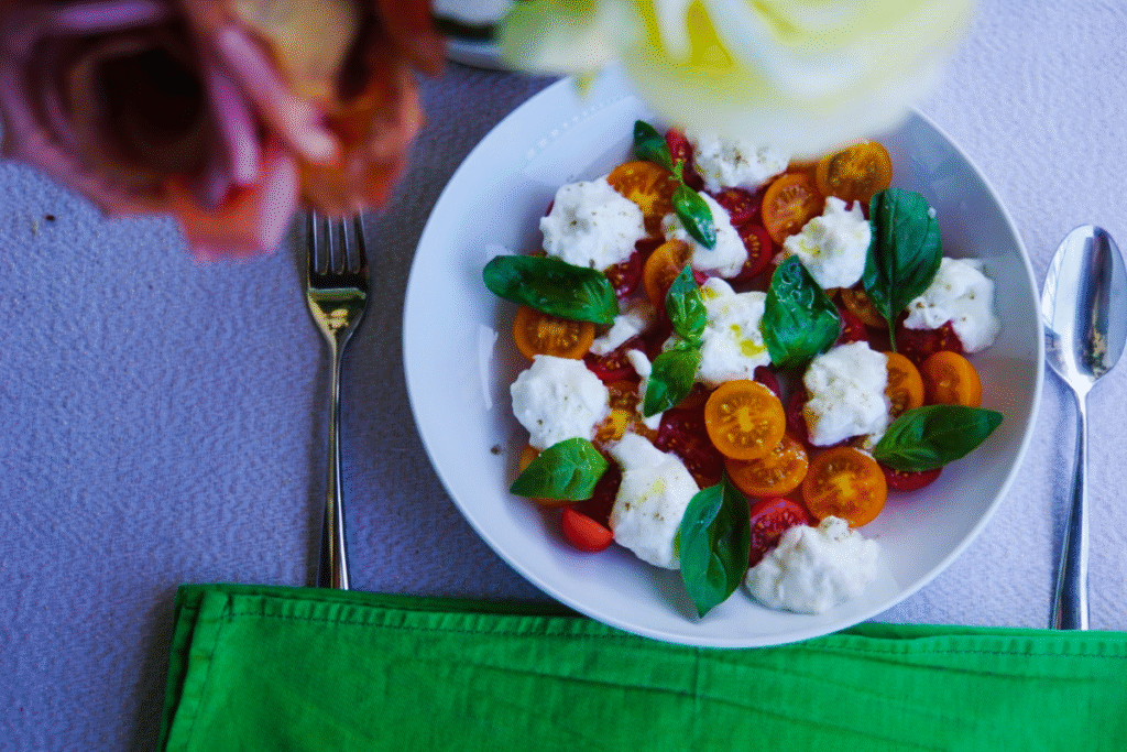 Burrata Caprese salad on a white plate with cherry tomatoes and basil, styled with a fork, spoon, and green cloth napkin on a table.