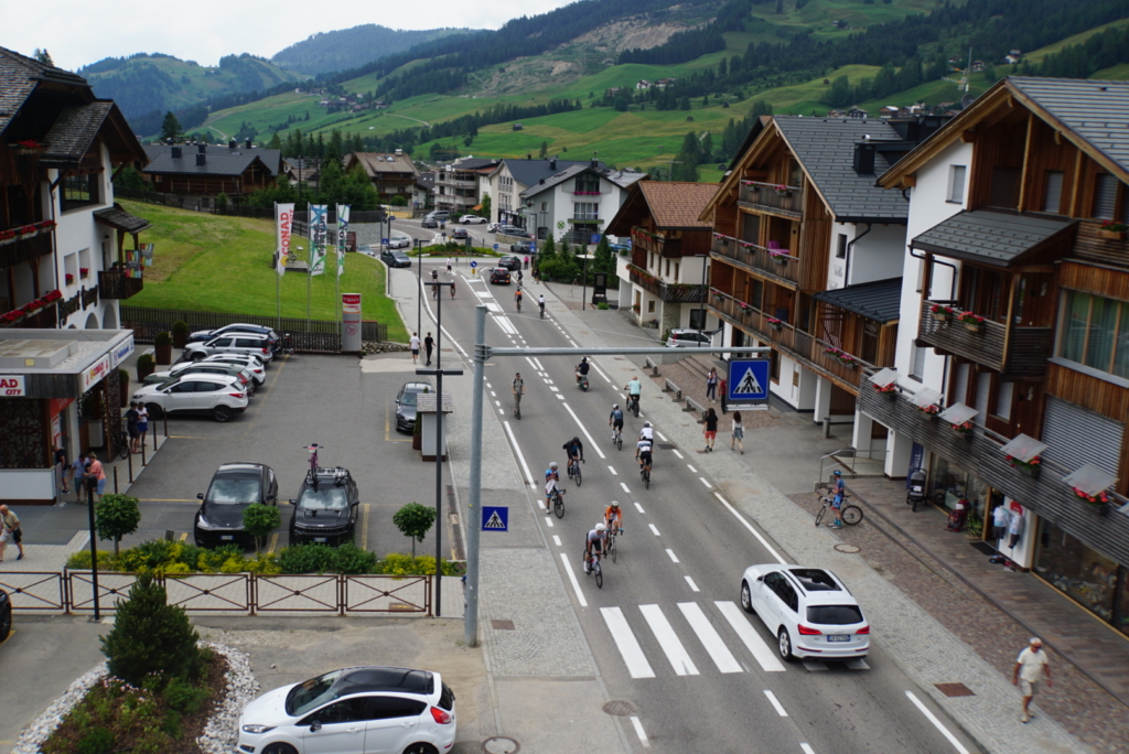 Cyclists riding through Corvara, Italy in summer, surrounded by Alpine-style buildings, green hills, and pedestrians enjoying the lively mountain town.