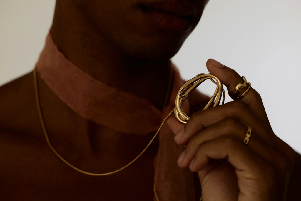Black woman in a dark brown dress, showcasing a thin gold necklace and matching rings.