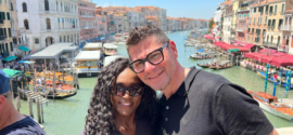 A couple standing on the Rialto Bridge in Venice, smiling in front of the Grand Canal lined with historic buildings and boats, on a bright summer day. This image captures a joyful moment that reflects the question: Is Venice worth visiting?