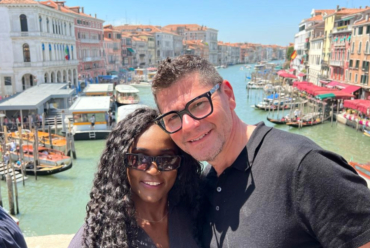 A couple standing on the Rialto Bridge in Venice, smiling in front of the Grand Canal lined with historic buildings and boats, on a bright summer day. This image captures a joyful moment that reflects the question: Is Venice worth visiting?
