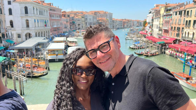 A couple standing on the Rialto Bridge in Venice, smiling in front of the Grand Canal lined with historic buildings and boats, on a bright summer day. This image captures a joyful moment that reflects the question: Is Venice worth visiting?