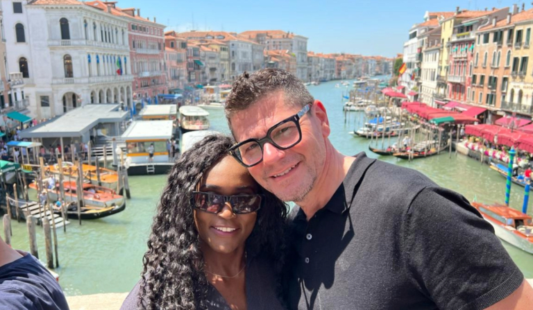 A couple standing on the Rialto Bridge in Venice, smiling in front of the Grand Canal lined with historic buildings and boats, on a bright summer day. This image captures a joyful moment that reflects the question: Is Venice worth visiting?