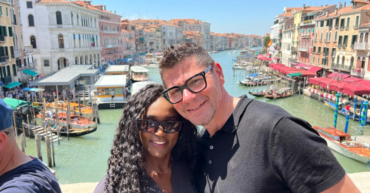 A couple standing on the Rialto Bridge in Venice, smiling in front of the Grand Canal lined with historic buildings and boats, on a bright summer day. This image captures a joyful moment that reflects the question: Is Venice worth visiting?