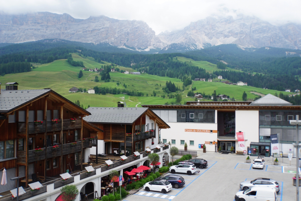 View of Corvara, Italy in summer showing Alpine buildings, café terraces, green hills, and Dolomite mountains in the background — one of the top reasons to visit Corvara in summer.