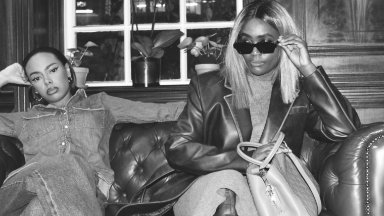 Two stylish women seated on a leather sofa, each showcasing distinct fashion personalities—one in double denim, the other in an oversized blazer and sunglasses.