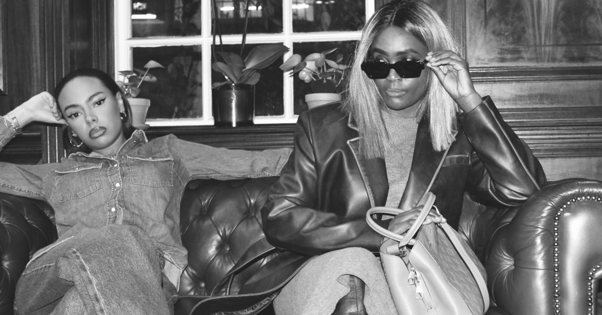 Two stylish women seated on a leather sofa, each showcasing distinct fashion personalities—one in double denim, the other in an oversized blazer and sunglasses.