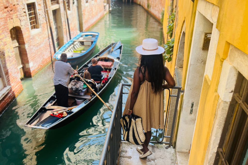 Woman with a panama hat on a balcony overlooking a Venice canal with a passing gondola.