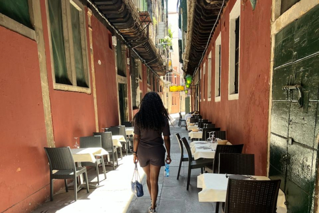 Woman walking in a narrow Venetian alley with outdoor restaurant tables.