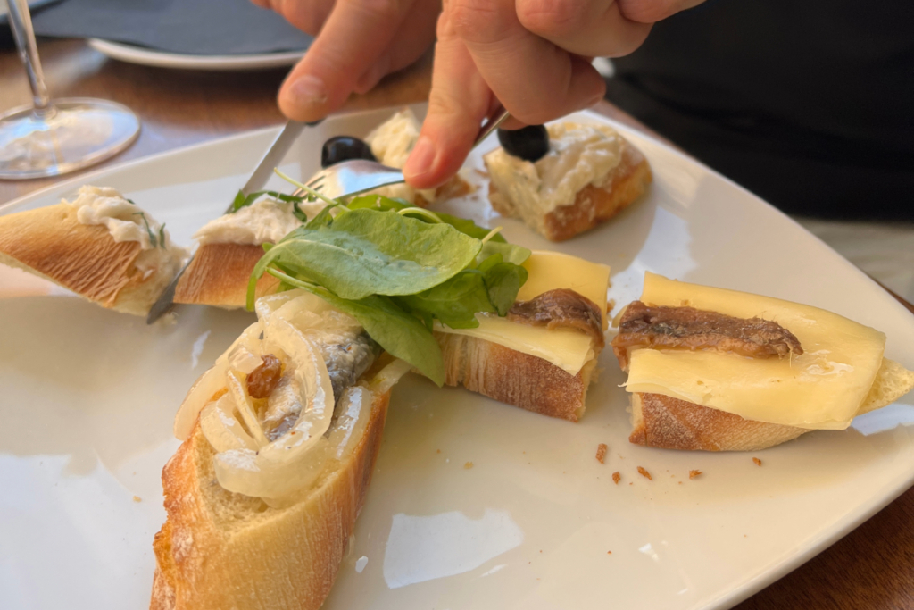 A close-up of hands cutting into a plate of assorted cicchetti, featuring toppings like onions, cheese, and a white spread.