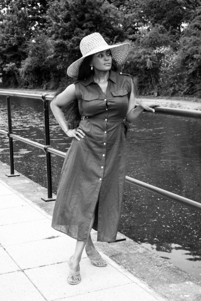 How to Accessorise an outfit: A woman in a sleeveless button-down dress and straw hat stands by a canal, showing a minimalist yet stylish summer look.