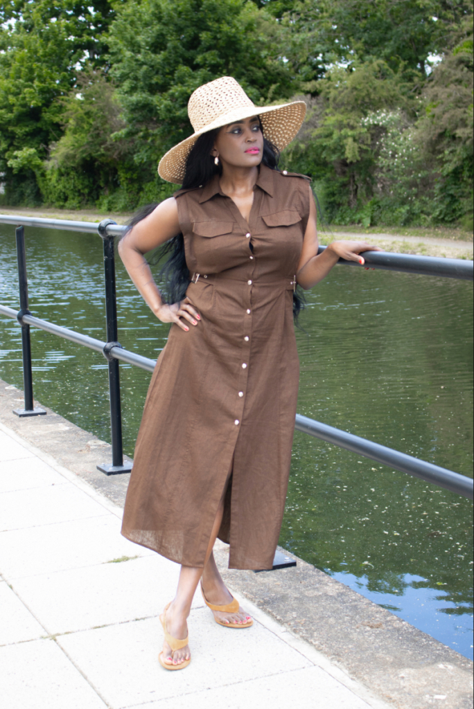 Elegant woman dressed for Sub-Saharan travel in July and August, wearing a brown linen cargo dress, wide-brim hat, and matching brown heeled sandals by a canal.