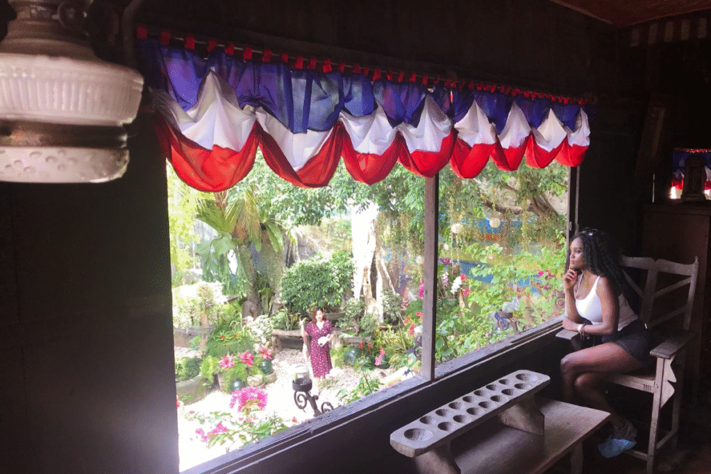 Black woman sitting pensively by a large window framed with red, white, and blue curtains, reflecting on the emotional toll of living abroad with a third world passport.