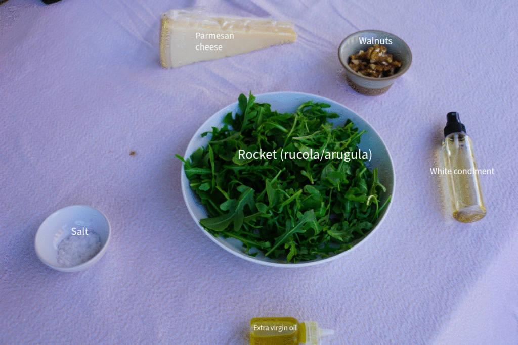 A collection of fresh rocket salad ingredients featuring a vibrant pile of rocket leaves, a large block of Parmigiano Reggiano cheese (Parmesan), walnut halves in a small bowl, a small bottle of extra virgin olive oil, and a white condiment.