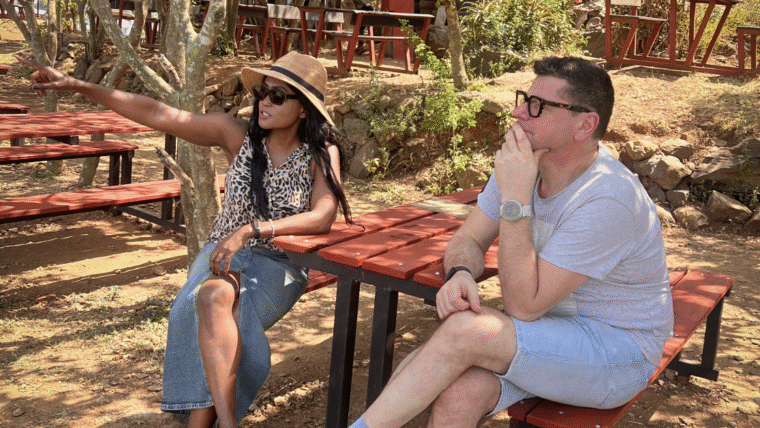 A woman sitting outdoors in sunglasses, pointing ahead while talking with a man, reflecting on her word of the year.