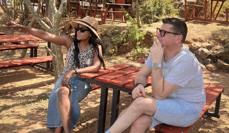 A woman sitting outdoors in sunglasses, pointing ahead while talking with a man, reflecting on her word of the year.