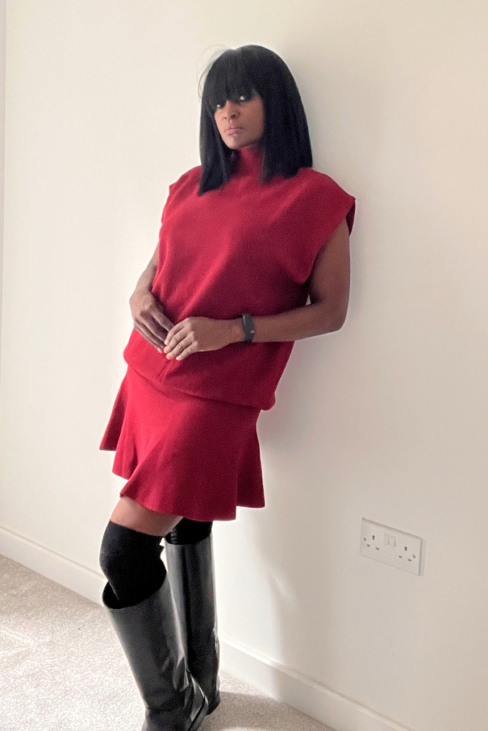 A woman demonstrates how to style a red knit top for a fall outfit, wearing a sleeveless red knit top, a matching short skirt, and black over-the-knee boots.
