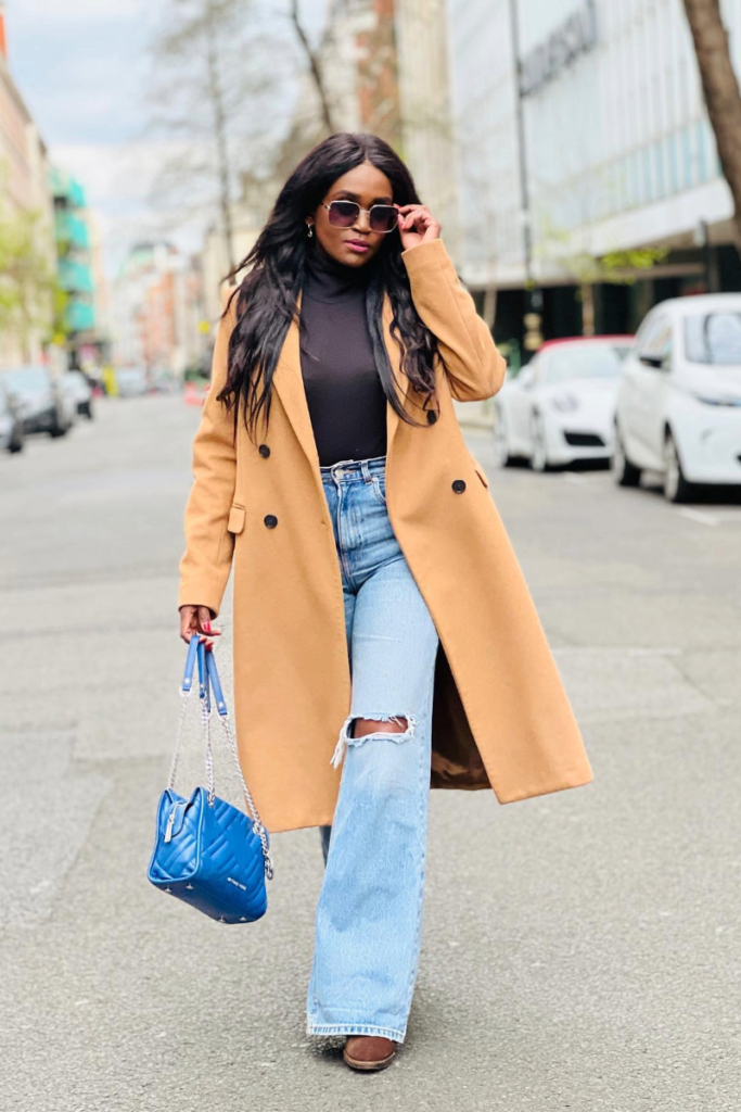 Stylish woman in camel coat, black turtleneck, and ripped flared jeans carrying a blue quilted handbag — showcasing must-have wardrobe essentials in a chic street-style look.