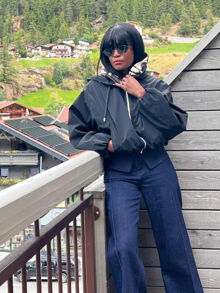 A woman standing on a wooden balcony in the Austrian Alps in September, wearing a black practical jacket, dark denim jeans, sunglasses, and a patterned scarf.