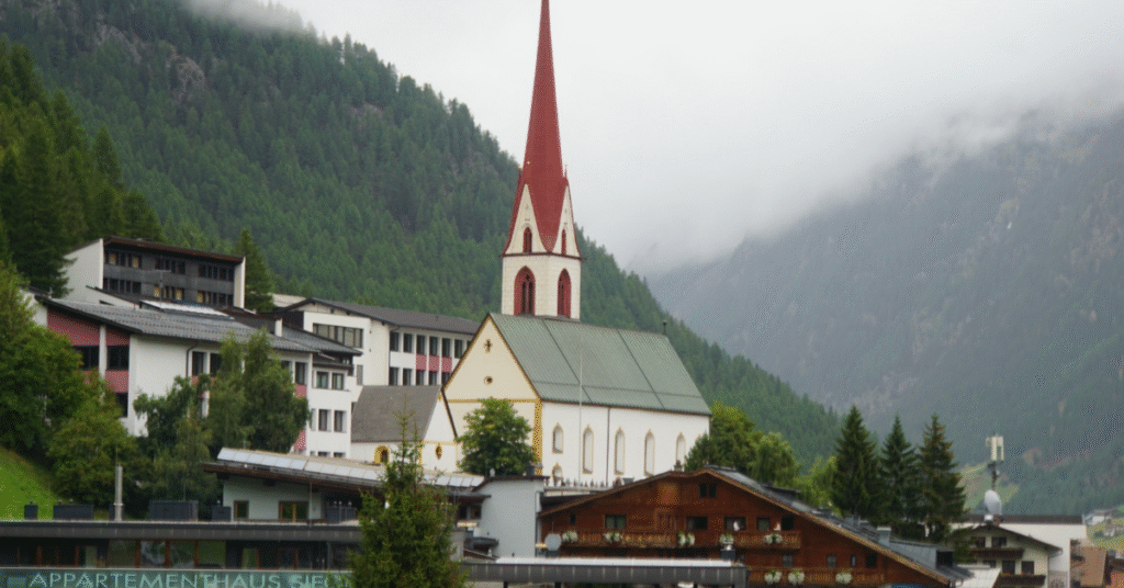 A white church with a red steeple stands in a Sölden village in late August, nestled in a valley surrounded by misty green mountains.