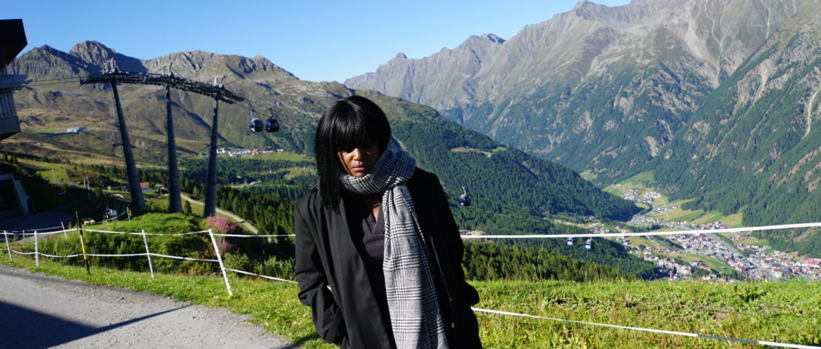 A woman stands on a mountain path in the Austrian Alps in September, wearing a long black coat and a large patterned scarf.
