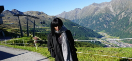 A woman stands on a mountain path in the Austrian Alps in September, wearing a long black coat and a large patterned scarf.