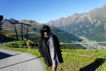 A woman stands on a mountain path in the Austrian Alps in September, wearing a long black coat and a large patterned scarf.