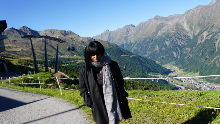 A woman stands on a mountain path in the Austrian Alps in September, wearing a long black coat and a large patterned scarf.