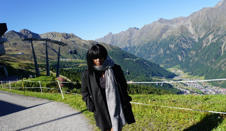 A woman stands on a mountain path in the Austrian Alps in September, wearing a long black coat and a large patterned scarf.