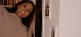 African woman smiling playfully at home beside framed photos, capturing a joyful moment as she prepares to celebrate Thanksgiving in her own way.