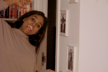 African woman smiling playfully at home beside framed photos, capturing a joyful moment as she prepares to celebrate Thanksgiving in her own way.