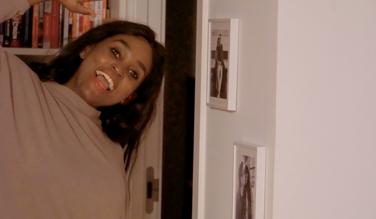 African woman smiling playfully at home beside framed photos, capturing a joyful moment as she prepares to celebrate Thanksgiving in her own way.