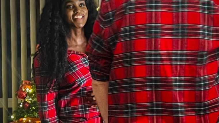 Woman wearing red tartan Christmas pyjamas at home beside a decorated Christmas tree, capturing a cosy festive moment indoors.
