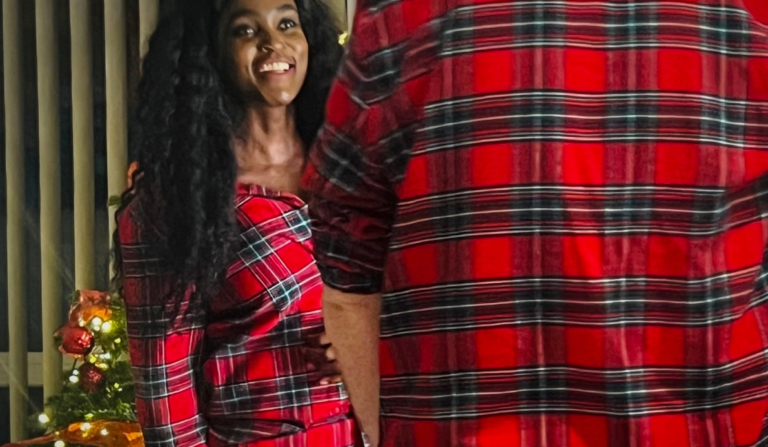 Woman wearing red tartan Christmas pyjamas at home beside a decorated Christmas tree, capturing a cosy festive moment indoors.